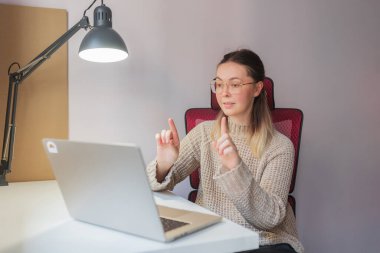Woman raising finger, appearing to explain something during video call.Confident gesture and serious look, young woman interacting via laptop, indoor lighting, red ergonomic chair and white workspace setup.