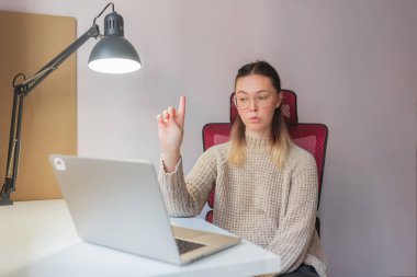 Woman raising finger, appearing to explain something during video call.Confident gesture and serious look, young woman interacting via laptop, indoor lighting, red ergonomic chair and white workspace setup.