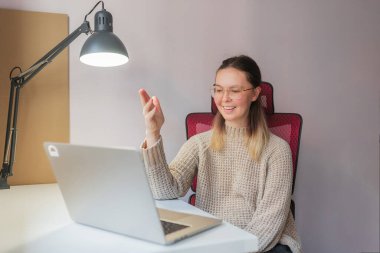 Genç bir kadın, evdeki video görüşmesi sırasında gülümsüyor ve el kol hareketi yapıyor. Bej kazaklı, dost canlısı bir kadın gümüş dizüstü bilgisayarda görüntülü sohbet yapıyor, kırmızı sandalye ve yumuşak masa lambası aydınlatması, çekici ve pozitif uzaktan haberleşme ayarları yapıyor..
