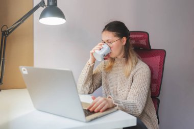 Laptopta çalışırken kupadan su içen bir kadın. Kırmızı örgü sandalye, bej örgü süveter, ve beyaz bardaktan yudumlarken odaklanmış bakışlar, temiz, modern beyaz masa başında oturan.