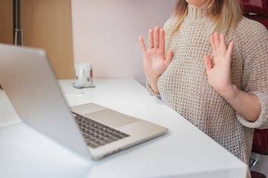 Online toplantı sırasında kadın ellerini hafifçe tarafsız bir şekilde kaldırır. Ellerini rahat bir şekilde kaldırır, sanal oturum sırasında masada oturur, açık renk kazak ve basit bilezik görünür, temiz bir masaüstü arka planı..