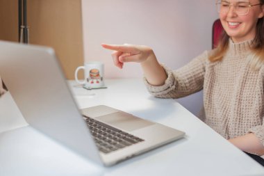 Neşeli ev videosu sırasında kadın gülümsüyor ve ekranda işaret ediyor. Parlak iç mekan sahnesinde gülümseyen kadın dizüstü bilgisayarı, günlük elbiseyi ve arka planda bardakla neşeli çevrimiçi etkileşimi işaret ediyor..