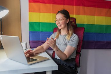 Gökkuşağı bayraklarıyla uzaktan yakından ilgilenen mutlu kadın hareketleri. Gri gömlekli, gözlüklü, gökkuşağı gurur bayrağı yanında oturan, parlak ev çalışma alanı ve dizüstü bilgisayar başında oturan, gri gömlekli, kendine güvenen genç kadın anlatıyor..