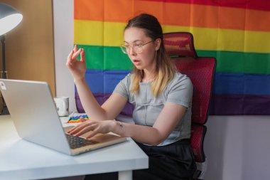 Kadın, uzaktan çağrı sırasında gökkuşağı bayrağıyla tepki verirken şaşkınlığını dile getiriyor. Arkasında gurur bayrağı bulunan parlak oda, kadın elini hafifçe kaldırıyor, ağzı açık şaşkınlık içinde, gündelik tişört ve ev ofis ortamını çevrimiçi toplantı sırasında gösteriyor..