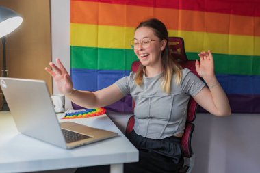 Happy woman greeting during video meeting, rainbow flag backdrop visible/Smiling young woman raising hand in front of laptop, sitting at desk with rainbow toy, pride-themed workspace.