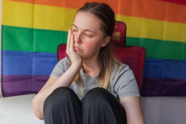 Düşünceli bir kadın, gökkuşağı gururunun önünde oturuyor. Dizleri çizilmiş, yüzü avuçlarında dinleniyor, düşünceli bir ifade, duvarda gurur bayrağı ve arkasında kırmızı bir sandalyeyle oturuyor.