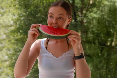 Gülümseyen kadın karpuz dilimlerini ısırır, beyaz bluz giyer, parlak yanaklar, siyah akıllı ceylan, bulanık yeşil arka plan, açık havada günlük saç stili, yaz mevsiminde meyve yiyerek neşeli ruh hali