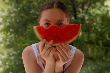 Genç bir kadın, elinde karpuz dilimi, yüzünde karpuz dilimi, yüzünde saklayan bir kadın, neşeli gözler, açık havada yaz tatili, üstsüz beyaz bluz, yeşil park arka planı..