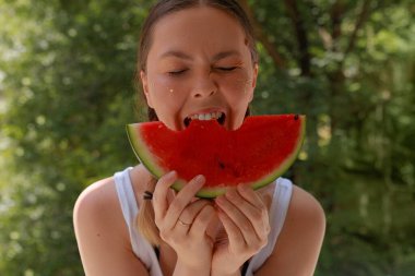 Karpuz dilimleyen genç bir kadın açık hava parkında gülümsüyor. Mutlu bir şekilde sulu karpuz dilimi ısıran bir kadın. Gözleri kapalı, parıltılı yanaklar, yaz kıyafeti, güneşli yeşil arka plan, park manzarası..