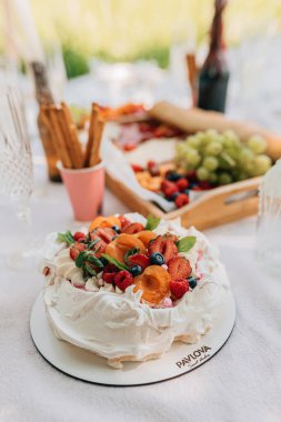 Dilimlenmiş kayısı, çilek, yabanmersini, ahududu, nane, pembe serpinti; şarap şişeleri, ekmek çubukları ve arka planda meyvelerle kaplı kremalı beyaz pavlova.
