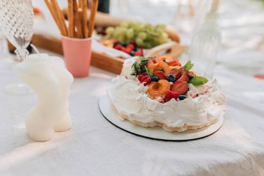 Fruit Pavlova keki dışarıda piknik atıştırmalıkları ve dekoratif detaylarla servis ediliyordu. Kremalı pasta, üzümler, meyveler, içki bardakları, kadınsı vücut şeklindeki mumlar ve yumuşak beyaz piknik örtüsü üzerinde gevrek ekmek çubukları ile hafif yaz sahnesi.
