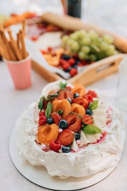 Piknik tedavisiyle çevrili, böğürtlen ve kayısıyla çırpılmış kremalı pavlova pastası. Kremanın zengin dokusuna, sulu meyvelere, nane süslemesine ve doğal ışık altında katmanlı tatlının üzerindeki pembe şeker toplarına odaklan.