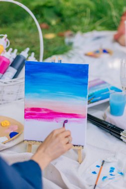 Fırça tuval üzerinde kayarak piknik boyama atölyesi sırasında pastel ufuk sesi yaratır. Çim, kumaş, boya malzemeleri ve içeceklerle çevrili bir sanatçı. Yakın çekim görünümlü yaz yaratıcılığı oturumu.