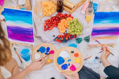 Açık hava pikniği sırasında çevrede aperatifler, kirazlar, üzümler ve meyve suyu bardaklarıyla canlı grafikleri gösteren iki tablo çiziliyor. Eller fırçaları ve paletleri parlak akrilik boyalarla tutar.