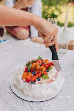 Açık hava kutlaması sırasında pasta dilimleme anının yakın çekimi. Yüzüklü el bıçak tutuyor, canlı meyve ve nane yapraklarıyla süslenmiş çırpılmış kremalı pasta servis etmeye hazırlanıyor. Piknik ayarları.