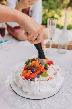 Gümüş yüzüklü el, kayısı, erik, yaban mersini ve nane ile süslenmiş bir kutlama pavlova pastasını keser. Krem şantili beyaz krema tabanı piknik sırasında şeffaf tabakta oturur..