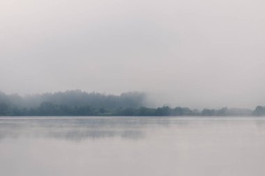Sisli orman çizgisi yoğun bulutlu gökyüzü altında geniş nehir boyunca yansıyor. Yoğun sabah sisi, sakin nehir yüzeyindeki yoğun ağaç çizgisi yansımasını yumuşatıyor, soluk renkler dingin, erken saatlerde el değmemiş doğal manzarayı anımsatıyor..