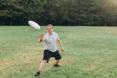 Konsantre ifadeye sahip atletik erkek, desenli beyaz gömlek, siyah şort, akıllı seğirme, çimenler üzerinde geniş adımlarla dinamik bir şekilde hareket ediyor, yumuşak doğal ışık altında tek elle yakalamaya hazır..