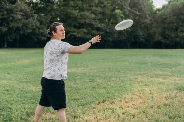 Odaklanmış beyaz gömlekli, siyah şortlu ve akıllı erkek, gün ışığında yumuşak orman zeminiyle çevrili geniş çimenli bir araziyle çevrili beyaz diski fırlatmak suretiyle takip ediyor..