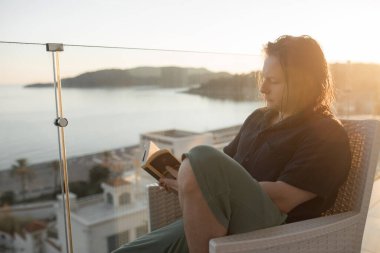 Islak saçlı, kitap okuyan rahat bir adam. Gün batımı sandalyede cam balkon. Deniz kıyısına bakan balkon. Adamın düşünceli yüz ifadesi. Modern şehir balkonunda yansıyan hafif ışık.