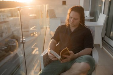 Altın saat boyunca ıslak saç okuma kitabı olan bir adam. Yalın ayak duruş, rattan sandalyeli şehir balkonunda oturuyor. Güneş ışığı, yüz hatlarını aydınlatıyor. 