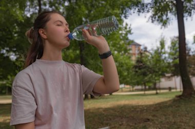 Soluk pembe gömlekli kadın, açık plastik şişeden su içiyor, gözleri kapalı, yeşil yapraklı park arkası, günlük kıyafetlerle dışarıda oturan kadın, gevşemiş su alma anı, yeniden kullanılabilir temiz şişe, doğal ışık., 