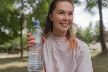 Yeşil ağaçların arasında dururken açık mavi şapkalı, temiz plastik bir su şişesi tutan kadın görülüyor. Düşük açı, dost ifadesini, yaprakların arasından süzülen doğal ışığı, yeşil arka planla çelişen soluk gömleği vurguluyor..