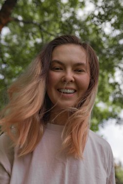 Direk bakışlı yakın portre, yumuşak saç meltemi, ılık doğal ışık gölgeleri, ağaçların derinlik kattığı sıradan yaşam tarzı..