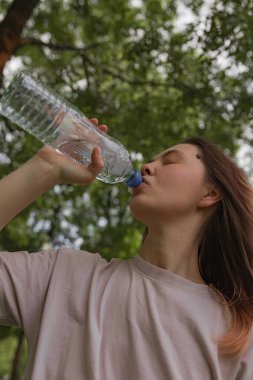 Genç bir kadının gözlerini kapatarak başını eğdiği açık havada yeşil ağaç dalları ve doğal ışıkla çevrili su içtiği odaklanmış bir görüntü. Ferahlık ve sağlığa vurgu yapar.