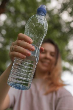 Kadın şişeyi ileri doğru uzatırken seçici odak noktası arka planda hafif bulanık bir yüz ve çevrede yeşillik. Doğal ışıklandırma suyu, sunma veya paylaşma jestini ön plana çıkarıyor.