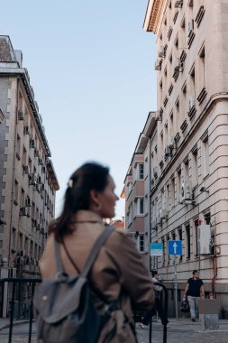 Sofya. Bulgaristan. 09.19.2025. Bej ceketli ve sırt çantalı bir kadının olduğu şehir yaşam tarzı, pencereli ve klimalı tarihi mimari ön cepheler, arka planda yayalar.
