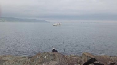 Glenarm, Kuzey İrlanda - 11 Ağustos 2025 Kuzey İrlanda 'nın kayalık kıyı şeridinde beyaz bir tekne kıyıda kalırken bir adam balık tutuyor. Sahne, çevresindeki deniz manzarası ile doğal gün ışığı altında kıyı balıkçılığı faaliyetlerini gösteriyor..