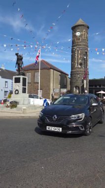 Bushmills, Kuzey İrlanda - 18 Ağustos 2025: Trafik güneşli bir öğleden sonra saat kulesi ve savaş anıtıyla köy merkezinden geçer