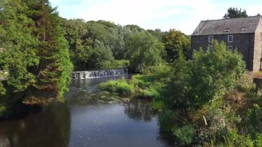 Bushmills, Kuzey İrlanda - 18 Ağustos 2025: Su, nehir kıyısındaki eski değirmen manzaralı, güneşli yaz günü Bush nehri boyunca akar