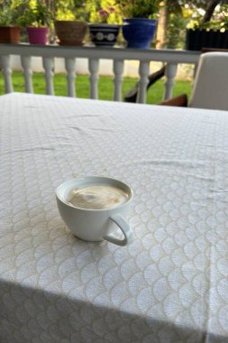 Masa örtüsüyle kaplı bir masanın üzerinde beyaz bir fincan kahve var. Masa çiçeklerle dolu bir bahçede duruyor..
