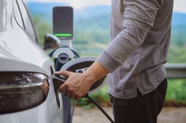 Elektrikli araba ya da ev istasyonda şarj oluyor. Adam beyaz güç kablosunu kullanıyor ve doğanın arka planına takıyor.