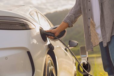 Elektrikli araba ya da ev istasyonda şarj oluyor. Adam beyaz güç kablosunu kullanıyor ve doğanın arka planına takıyor..