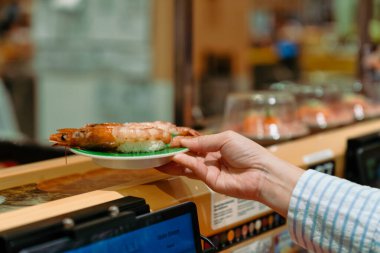 Modern bir restorandaki taşıma bandından taze suşi tabağı alan bir el resmi. Otomatik suşi servisinin rahatlığı, tazeliği ve eşsiz yemek deneyimi..