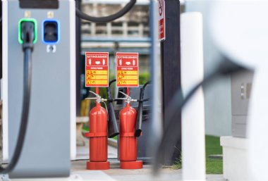 Yukarıda güvenlik talimatları bulunan EV şarj istasyonunda iki kırmızı yangın söndürücünün yakın çekimi, acil durum hazırlığını sağlamak ve modern elektrikli araç altyapısında yangın güvenliği tedbirlerini vurgulamak.