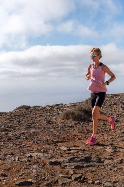 Pembe ve siyah atletik giysiler içindeki kadın koşucu, yukarıda açık bir gökyüzü olan kayalık bir yolda güvenli adımlarla yürüyor, dayanıklılığını ve açık hava sporlarına olan tutkusunu gösteriyor..