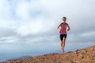 Güneş gözlüklü bir kadın bulutlu bir gökyüzünün altında dağ yolunda koşuyor. Fitness, kararlılık ve açık hava aktivitesi.