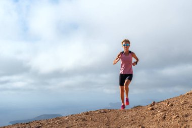Bulutlu bir gökyüzünün altında koşan atletik giysiler giyen bir kadın doğadaki uygunluğu ve kararlılığı temsil ediyor..