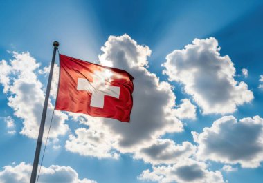 İsviçre bayrağı, beyaz haçlı kırmızı bir meydan, rüzgarda dalgalanıyor çarpıcı bulutlar ve güneş ışınlarıyla dolu canlı mavi bir gökyüzüne karşı.