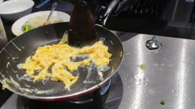Şef çırpılmış yumurtaları ocakta tavada karıştırıyor. Yakın plan çekimler, altın kabarık dokuyu vurguluyor. Hızlı bir kahvaltı hazırlamak için bir aşçılık gösterisi.