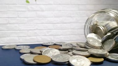 Mavi bir yüzey üzerinde eğimli cam bir kavanozun etrafına saçılmış paraların statik yakın çekim videosu beyaz tuğlaların arka planına karşı. Bu kompozisyon tasarruf, finans ve mali konuları çağrıştırır