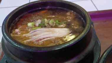 Taş kaselerde servis edilen buharı tüten Kore sıcak tencere koleksiyonunun yakın çekim görüntüsü. Taze sebzeler, mantarlar ve dilimlenmiş taze soğan gibi narin süslemelerle dolu zengin çorbalar.