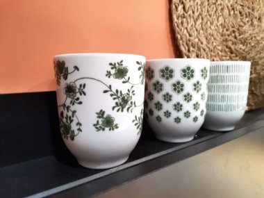 Değişen yeşil desenleri, çiçekli sarmaşıkları, geometrik çiçekleri ve doğrusal tasarımları olan beyaz bardaklardan oluşan bir üçlü zarif bir şekilde bir rafa dizilir..