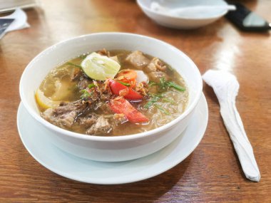 Taze limon ve domates garnitürüyle süslenmiş Soto Mie çorba kasesinin büyüleyici görüntüsü ahşap bir masanın üzerinde. Tek bir çerçevede yakalanan aromatik bir mutfak deneyimi..