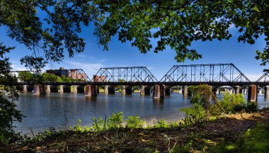 Nehirden geçen eski çelik köprü. Köprü ön planla çerçevelenmiş ve açık mavi bir gökyüzü olan ağaçlar arkaplan gibi..