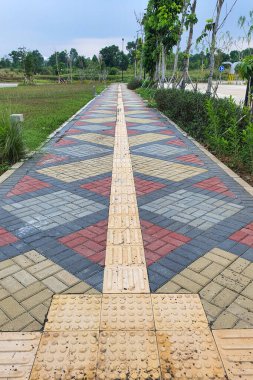 pedestrian pathway with a colorful paving block pattern and guiding block winds through a park, lined with lush green grass and trees. clipart